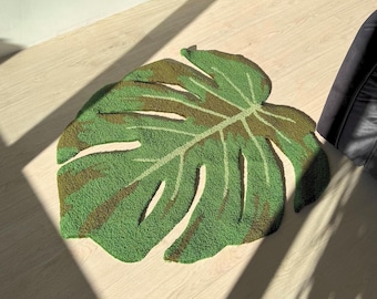 Op de afbeelding: Een getuft tapijt in de vorm van een groot monstera-blad, met een getextureerd oppervlak in een levendig groen en donkerbruin kleurpatroon. Het tapijt heeft diepe uitsparingen langs de randen om het natuurlijke uiterlijk na te bootsen.