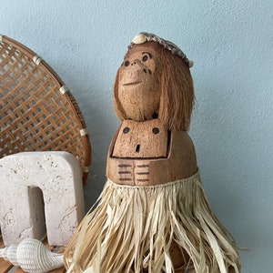 May include: A coconut monkey figurine with a straw skirt and a shell crown. The figurine is sitting on a wooden shelf with books.