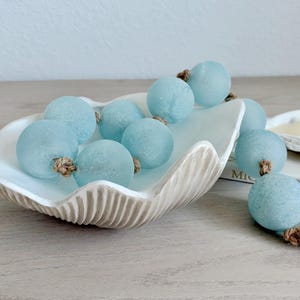 May include: A white ceramic bowl shaped like a seashell containing a strand of light blue, frosted glass spheres strung together with brown jute twine. The strand of beads extends out of the bowl and rests on the surface next to it.