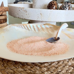 May include: A large, iridescent white seashell bowl filled with fine pink salt, accompanied by a small, curved spoon with a white handle and a dark metal bowl, resting on a woven circular mat.