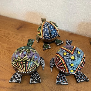 Könnte beinhalten: Drei Holzschildkrötenfiguren mit farbenfrohen Punktmustern. Die Schildkröten haben schwarze Beine und Köpfe, und die Panzer sind mit blauen, gelben, roten und lila Punkten verziert. Sie werden auf einer Holzoberfläche ausgestellt.