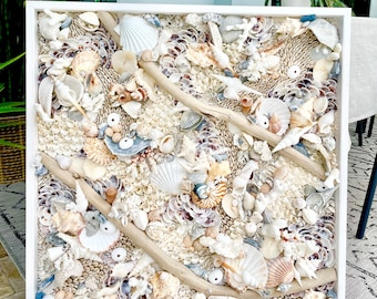 May include: A white framed square wall art piece made of various seashells, driftwood, and other natural elements. The piece has a beachy and coastal theme.