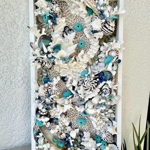 May include: A rectangular wall art piece featuring a collage of seashells, coral, and sea urchins in a white frame. The artwork incorporates various textures and colors, including white, teal, and shades of blue and gray. The composition is dense and detailed.
