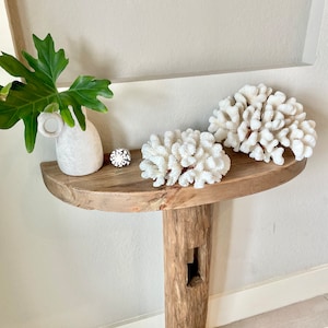 K&ouml;nnte beinhalten: Ein rustikaler Holz-Konsolentisch mit einem einzigen, dicken Holzbein. Der Tisch ist mit wei&szlig;em Korallen und einer wei&szlig;en Keramikvase mit einer gr&uuml;nen Pflanze dekoriert.