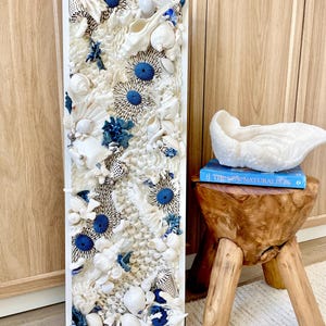 May include: A rectangular white framed wall art piece featuring an intricate arrangement of seashells, sea urchins, and coral in shades of white and blue. The artwork is displayed near a wooden stool with a decorative coral sculpture and a book.