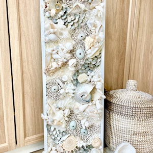 May include: A rectangular wall art piece featuring a collage of various seashells in shades of white, cream, and pale blue. The shells are arranged in a textured, three-dimensional pattern within a white frame. The artwork evokes a coastal or nautical theme.
