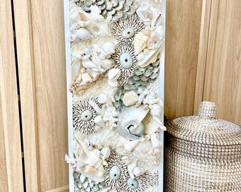 May include: A rectangular wall art piece featuring a collage of various seashells in shades of white, cream, and pale blue. The shells are arranged in a textured, three-dimensional pattern within a white frame. The artwork evokes a coastal or nautical theme.