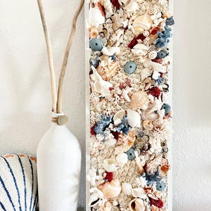 May include: A rectangular wall art piece featuring a collage of seashells, sea urchins, and coral in shades of white, beige, red, and blue. The artwork is framed in white and evokes a coastal theme. A white vase with driftwood is next to the art.