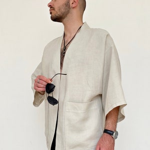May include: A light beige linen kimono with a single patch pocket on the left side. The kimono has a long, open front and wide sleeves. The model is wearing the kimono with a black necklace and sunglasses.