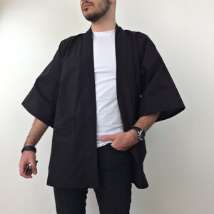 May include: A black kimono with long sleeves and a pocket on the left side. The kimono is open in the front and is worn over a white t-shirt and black jeans.