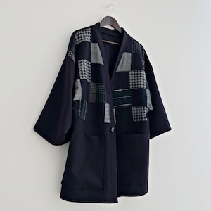 Może przedstawiać: Czarna kurtka z długimi rękawami z patchworkowym wzorem z przodu. Patchwork wykonany jest z różnych tkanin w odcieniach szarości i czerni. Kurtka zapinana jest na guziki i ma dwie kieszenie.