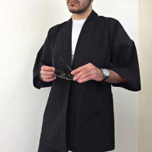 May include: A black kimono-style jacket with long sleeves and a simple design. The jacket has two pockets on the front and is made of a lightweight fabric.
