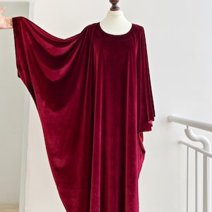 May include: A long, flowing, burgundy velvet dress with a round neckline and wide sleeves. The dress is displayed on a mannequin against a white background. The fabric drapes elegantly, showcasing the dress's luxurious texture and design.