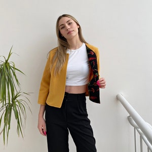 May include: A mustard yellow cropped jacket with a black and red floral print lining. The jacket has a collar and long sleeves.