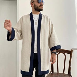 May include: A beige linen kimono-style jacket with navy blue trim. The jacket has a relaxed fit and three-quarter length sleeves. The navy blue trim is featured on the lapels, cuffs, and hem. The jacket is worn over a white t-shirt and black pants.