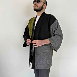 May include: A color-blocked kimono-style jacket in black, olive green, and gray. The jacket features a shawl collar and wide sleeves. The garment is made of a textured fabric and has a relaxed fit. The jacket has two front pockets.