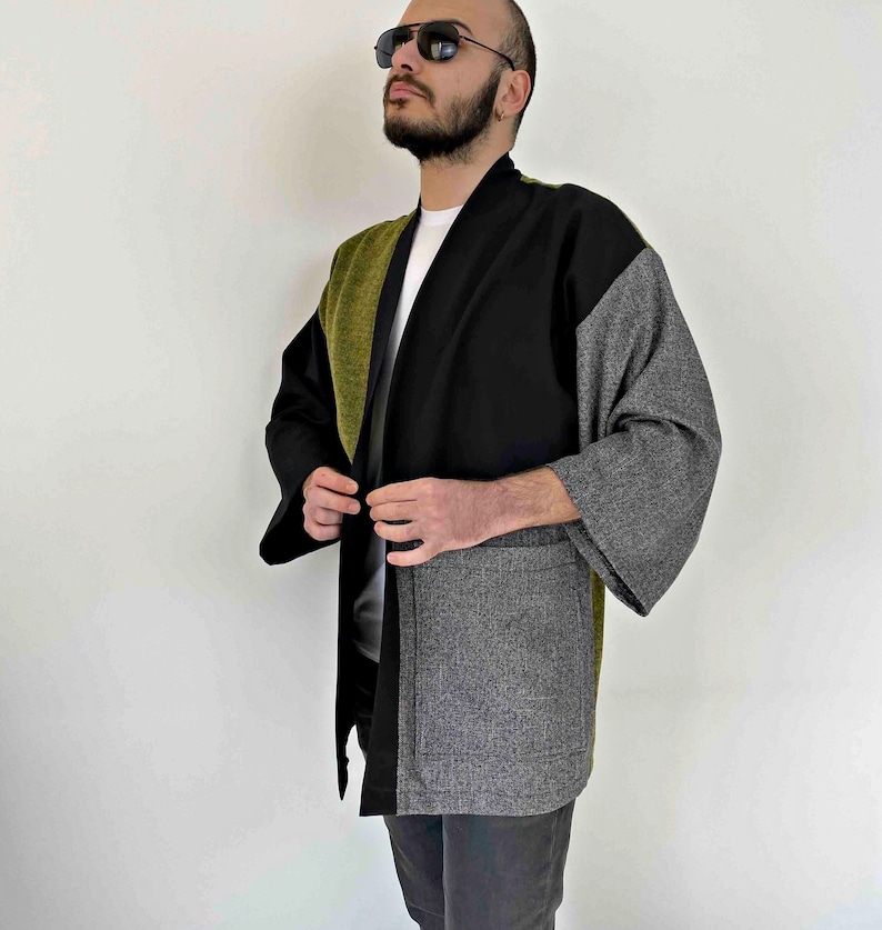May include: A color-blocked kimono-style jacket in black, olive green, and gray. The jacket features a shawl collar and wide sleeves. The garment is made of a textured fabric and has a relaxed fit. The jacket has two front pockets.