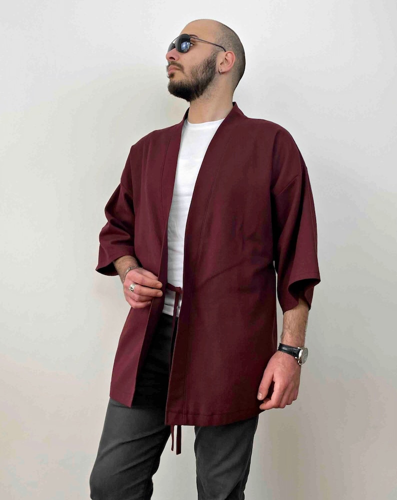 An adult male model wearing a plain burgundy color kimono. The Haori's length is hanging down around the man's buttocks. This kimono jacket is collarless, has lacings down around the model's belly and it has 3/4, wide sleeves.