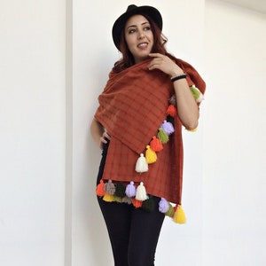 May include: A rust-colored woven shawl with colorful tassels. The shawl is made of a soft, lightweight fabric and has a checkered pattern. The tassels are made of yarn in various colors, including orange, yellow, green, purple, and white.