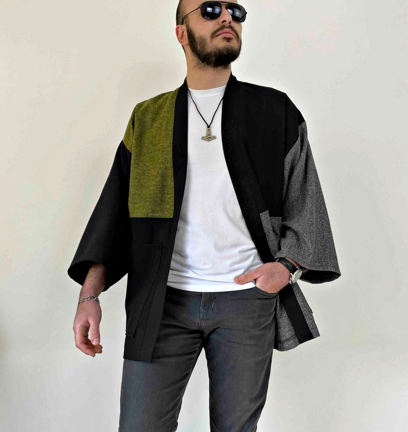 May include: A patchwork kimono-style jacket in black, olive green, and gray. The jacket has wide sleeves and a relaxed fit. The model is wearing a white t-shirt and dark gray pants. The jacket features a unique color-block design.