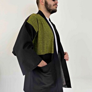 May include: A black and olive green kimono-style jacket with large pockets. The jacket has a contrasting olive green panel across the upper chest and shoulders. The sleeves are wide and the jacket is open in the front.