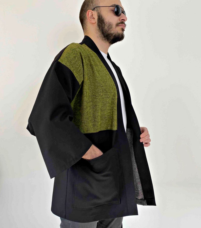 May include: A black and olive green kimono-style jacket with large pockets. The jacket has a contrasting olive green panel across the upper chest and shoulders. The sleeves are wide and the jacket is open in the front.