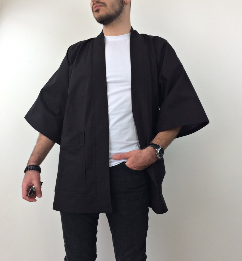 May include: A black kimono with long sleeves and a pocket on the left side. The kimono is open in the front and is worn over a white t-shirt and black jeans.