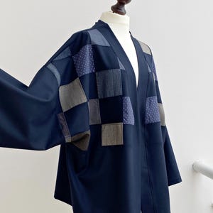 May include: A navy blue patchwork jacket with a relaxed fit. The jacket has a V-neck and long sleeves. The patchwork design incorporates squares of various textured fabrics in shades of blue and grey, creating a unique and stylish garment.