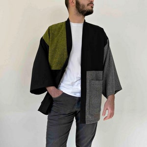 May include: A patchwork kimono-style jacket in black, olive green, and gray. The jacket has wide sleeves and a relaxed fit. The front features a pocket on the right side. The model is wearing a white t-shirt and gray pants.