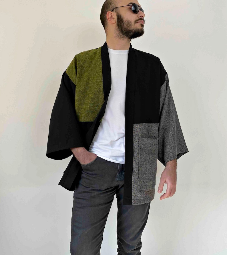 May include: A patchwork kimono-style jacket in black, olive green, and gray. The jacket has wide sleeves and a relaxed fit. The front features a pocket on the right side. The model is wearing a white t-shirt and gray pants.