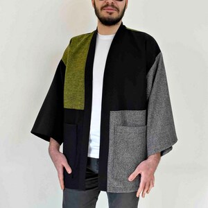 May include: A patchwork kimono-style jacket in black, olive green, and gray. The jacket features large pockets and a relaxed fit. The sleeves are wide and the garment is open in the front.