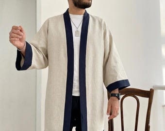 May include: A beige open-front kimono-style jacket featuring contrasting dark navy blue trim along the collar, front opening, and sleeve cuffs. The garment has wide, three-quarter length sleeves and a straight hemline, worn over a white t-shirt.