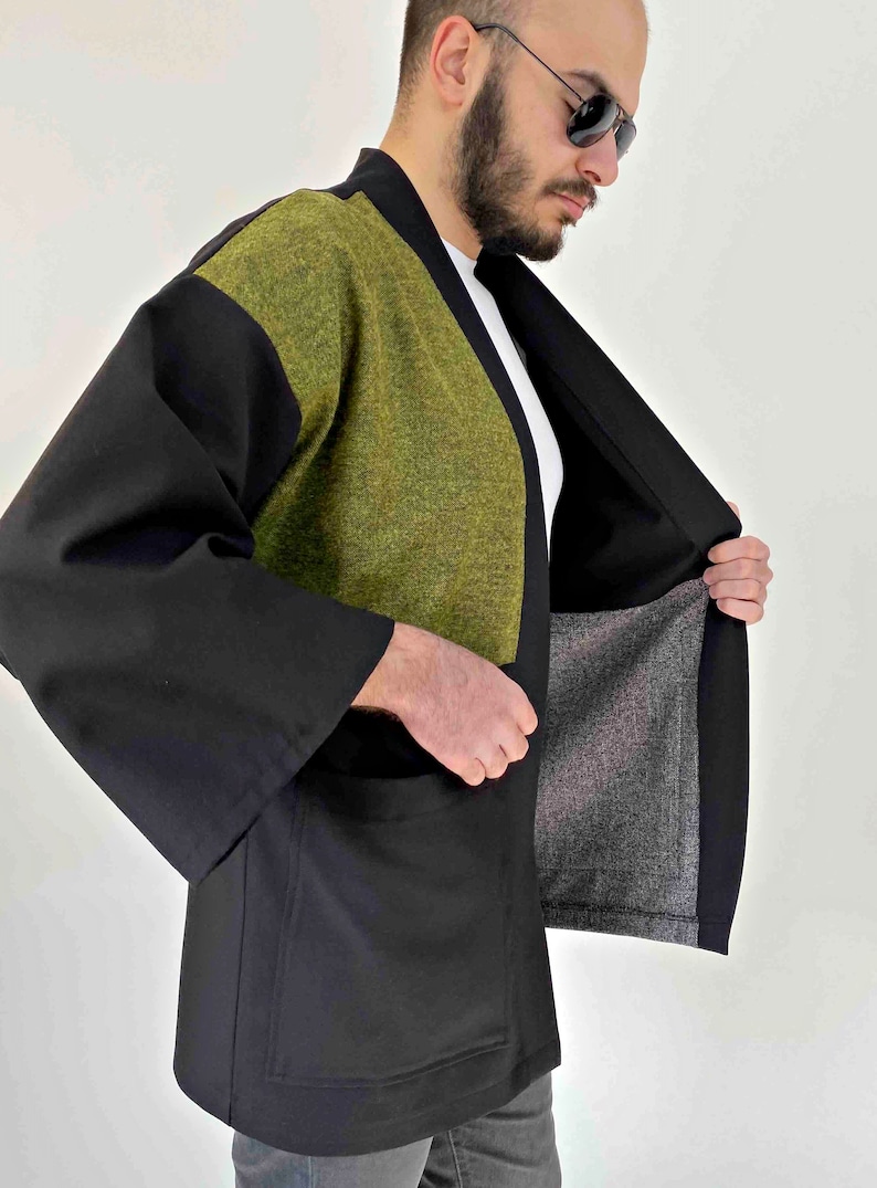 May include: A black kimono-style jacket with a green textured panel on the upper chest and shoulders. The jacket has wide sleeves and a relaxed fit. The interior lining is a gray fabric.