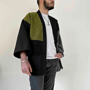 May include: A black, olive green, and gray kimono-style jacket with wide sleeves and a relaxed fit. The jacket features a color-block design with a large olive green panel on the upper chest and back. The jacket has two front pockets.