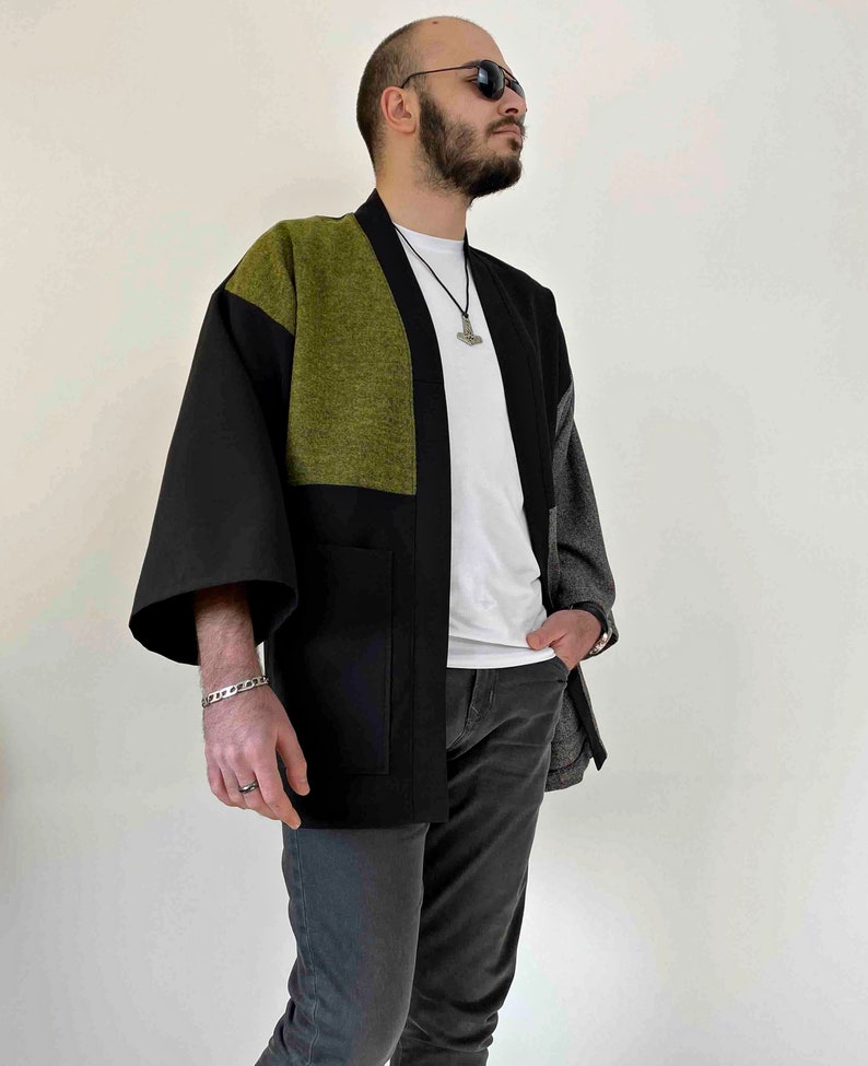 May include: A black, olive green, and gray kimono-style jacket with wide sleeves and a relaxed fit. The jacket features a color-block design with a large olive green panel on the upper chest and back. The jacket has two front pockets.