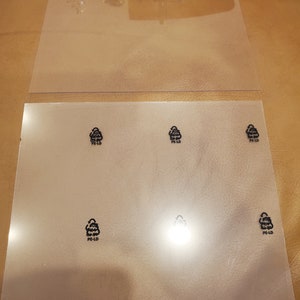 Könnte beinhalten: Zwei transparente Kunststofffolien, eine über der anderen. Die untere Folie hat sechs Recycling-Symbole mit dem Text "PE-LD" darunter. Die Folien liegen auf einer braunen Oberfläche.