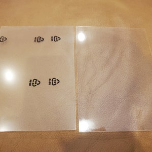 Könnte beinhalten: Zwei transparente, rechteckige Kunststoffplatten. Auf einer Platte sind mehrere schwarze Recycling-Symbole aufgedruckt. Die Platten liegen auf einer braunen, lederartigen Oberfläche.