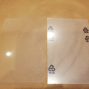 Könnte beinhalten: Zwei transparente, rechteckige Kunststoffplatten mit abgerundeten Ecken. Eine Platte ist glatt, die andere hat Recycling-Symbole und den Text "PE-LD" und "04". Die Platten liegen auf einer beigen Oberfläche.