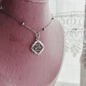 Può includere: Una collana d'argento con un ciondolo d'argento che rappresenta il simbolo del chakra Anahata. Il ciondolo è di forma quadrata con un cuore al centro. La collana è su un espositore in velluto rosa.