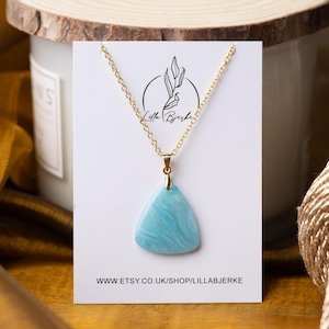 Puede incluir: Un collar de cadena dorada con un colgante triangular azul claro. El colgante tiene un patrón de mármol. El collar se muestra en una tarjeta blanca con el texto "Little Birdie" y una dirección de sitio web.