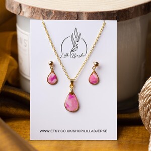 Puede incluir: Un collar con un colgante de gota de agua con patrón de mármol rosa y blanco y pendientes a juego con detalles dorados. Las joyas se muestran en una tarjeta blanca con el texto "Lilla Bjerke" y una dirección de sitio web.