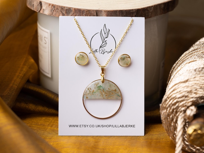 Puede incluir: Un conjunto de collar y pendientes de oro con un colgante redondo con un dise&ntilde;o de resina verde y dorada. El colgante es medio c&iacute;rculo con un marco de alambre dorado. Los pendientes son peque&ntilde;os y redondos con un dise&ntilde;o similar de resina verde y dorada. Las joyas se muestran en una tarjeta blanca con el texto "Lilla Bj&ouml;rke" y "www.etsy.co.uk/shop/lillabj&ouml;rke".