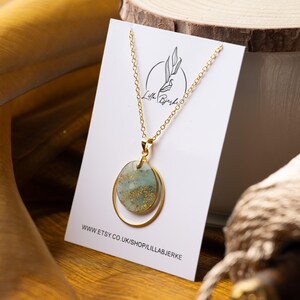 Puede incluir: Collar de oro con un colgante verde y dorado con dos círculos superpuestos. El colgante está sobre una tarjeta blanca con el texto "Lilla Björke" y "www.etsy.co.uk/shop/lillabjörke".