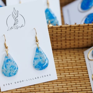 Puede incluir: Un par de pendientes de gota azul y blanco con ganchos dorados. Los pendientes se muestran en una tarjeta blanca con el texto "Lilla Bjerke" y "Etsy Shop: Lillabjerke".