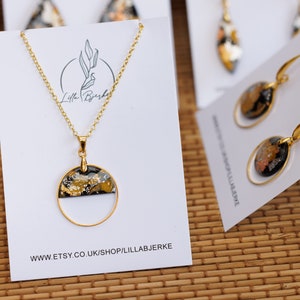 Puede incluir: Un conjunto de collar y pendientes de oro con colgantes de resina negra y dorada. Los colgantes son circulares con un borde dorado y un fondo negro con motas doradas. El collar tiene una cadena de oro delicada y los pendientes tienen ganchos de oro. La joyería se muestra en una tarjeta blanca con el texto "Lilla Bjerke" y "www.etsy.co.uk/shop/lillabjerke".