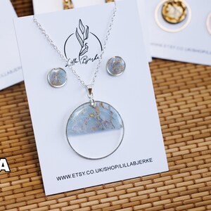 Puede incluir: Un collar de plata con un gran colgante redondo con un dise&ntilde;o marmoleado azul y dorado. El colgante est&aacute; engastado en una resina transparente. El collar viene con un par de pendientes a juego.