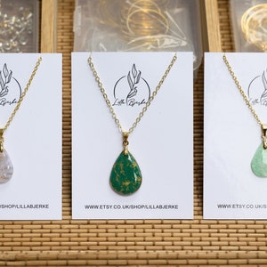 Puede incluir: Tres collares de oro con colgantes en forma de lágrima sobre tarjetas blancas. Los colgantes están hechos de resina y tienen diferentes colores y patrones: blanco con motas doradas, verde con motas doradas y verde claro. Las tarjetas tienen el texto "Lilla Bjerke" y "www.etsy.co.uk/shop/lillabjerke".