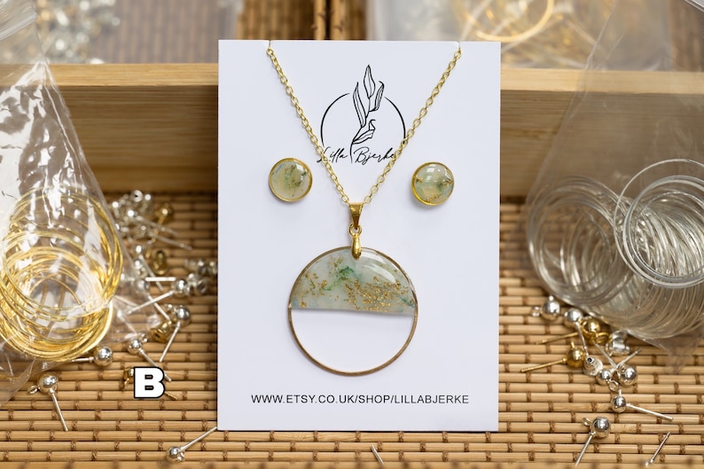 Puede incluir: Un conjunto de collar y pendientes de oro con un colgante redondo que presenta un dise&ntilde;o de resina verde y blanca con motas doradas. El colgante est&aacute; en una cadena de oro. Los pendientes son peque&ntilde;os y redondos con un dise&ntilde;o similar. El conjunto se muestra en una tarjeta blanca con el texto "Lilla Bj&ouml;rke" y "WWW.ETSY.CO.UK/SHOP/LILLABJ&Ouml;RKE".