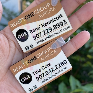 May include: Two brown and white business cards with black text and a black circle with the word "ONE" in white. The top card has the text "REALTYONEGROUP AURORA ONE Rene' Kennicott 907.229.8993 rene@renekennicott.com". The bottom card has the text "REALTYONEGROUP AURORA ONE Tina Cole 907.242.2280 tcole220@gmail.com".