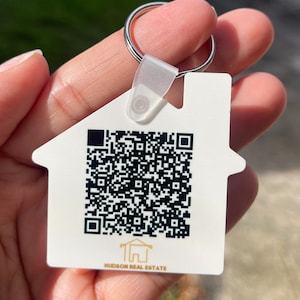 May include: White house-shaped keychain with a black and white QR code and the text "HUDSON REAL ESTATE" in gold lettering.
